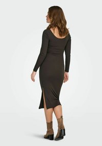 Abito midi a maniche lunghe in maglia a coste di colore verde scuro con spacco laterale. Presenta una schiena a scollo ampio e un design aderente. Indossato con stivaletti marroni.