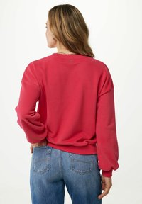 Sudadera de algodón roja con hombros caídos y puños acanalados. Presenta una pequeña etiqueta de marca cerca del cuello y un corte relajado. Se lleva con jeans azules.