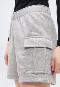 Κοντινή λήψη ανοιχτόγκρι καθημερινών σορτς με μεγάλη πλαϊνή τσέπη και καπάκι, φορεμένα με μαύρο μακρυμάνικο τοπ.