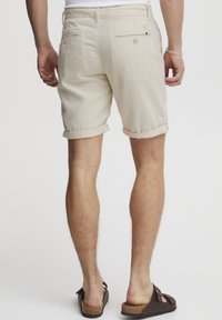 Beige lenvászon rövidnadrág, felgöngyölített szárakkal, hátul gombbal záródó zsebbel, barna slip-on szandálokkal viselve.