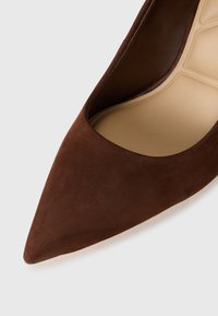 Brun semsket høyhælte sko med spiss tå og utskjæring på siden, med en glatt beige såle og subtil tekstur.