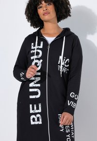 Zwarte zip-up hoodie jurk met witte tekstgraphics, voorzien van een capuchon, lange mouwen en zijzakken. Gemaakt van een zachte, comfortabele stof.