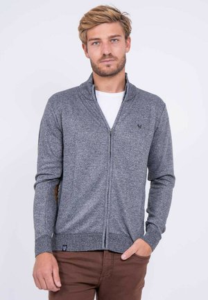 Homme aux cheveux blonds portant un pull gris zippé sur une chemise blanche et un pantalon marron, debout sur un fond blanc uni.