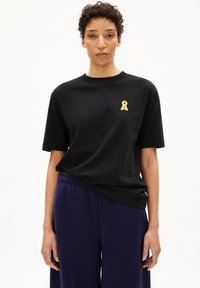 Zwart T-shirt met een gele lintontwerp op de linkerkant van de borst, gemaakt van zachte stof. Gecombineerd met loszittende marineblauwe broek.