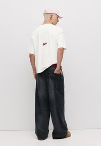 Hvid oversized t-shirt med rødt logo på ryggen, parret med løse mørke denimjeans og brune støvler. Lyserød kasket båret bagfra.