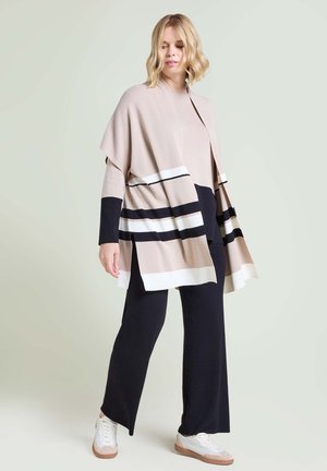 Woman wearing beige, black, and white striped poncho over black wide-leg pants and white sneakers, standing against a light background.