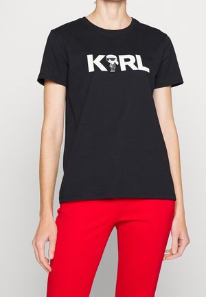 Personne portant un t-shirt noir avec un texte blanc "KARL" et un personnage de dessin animé, associé à un pantalon rouge vif sur un fond gris clair.
