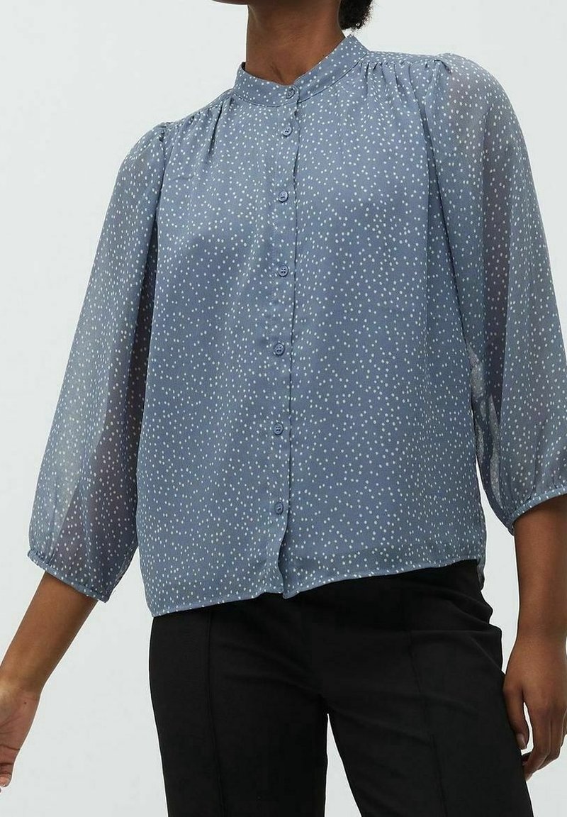 Blouse bleu clair en tissu transparent avec des pois blancs, boutonnage sur le devant, col officier et manches bouffantes trois-quarts.