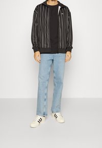 Svart zip-up träningsjacka med vita vertikala ränder, kombinerad med en svart t-shirt och ljusblå raka jeans. Vita sneakers med svarta ränder.