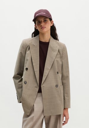 Woman wearing beige double-breasted blazer and matching pants, dark brown shirt, and maroon cap with white logo, standing against plain background.