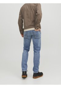 Brun jacka med knappdetaljer, krämfärgad tröja under, blå jeans med bakfickor, svarta skor med bruna sulor, vy bakifrån.