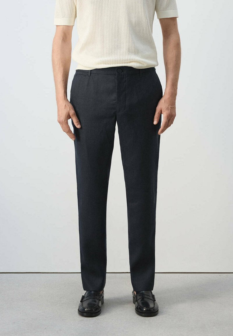 Homme portant un pantalon noir, des mocassins noirs, et une chemise tricotée blanche à manches courtes, debout devant un fond clair uni.