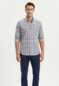 Hombre vistiendo una camisa de cuadros multicolores con las mangas arremangadas y pantalones oscuros, de pie contra un fondo liso.