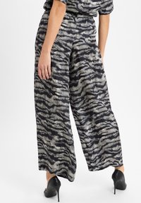 Vrouw in zwart-grijze zebraprint wijde broek en zwarte hoge hakken, staand tegen een effen achtergrond.