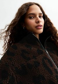 Bruine fleece zip-up jas met een luipaardpatroon, met een hoge kraag en een zachte textuur. Het model heeft golvend haar en zilveren ringoorbellen.