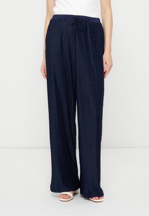 Γυναίκα με φαρδιές, ναυτικές μπλε υφασμάτινες παντελόνες με κορδόνι στη μέση και λευκά σανδάλια με ανοιχτά δάχτυλα, στέκεται μπροστά σε απλό φωτεινό φόντο.