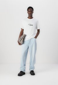 Camiseta blanca con el logo "JACK & JONES", vaqueros azul claro y zapatos negros gruesos. Sosteniendo una bolsa gris. Fondo liso.