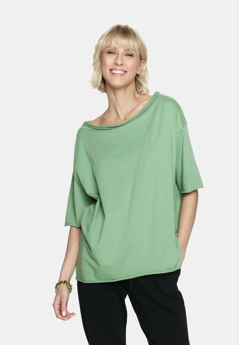 Luźna zielona bluzka z miękkiego materiału, z szerokim dekoltem i krótkimi rękawami, zestawiona z czarnymi spodniami.