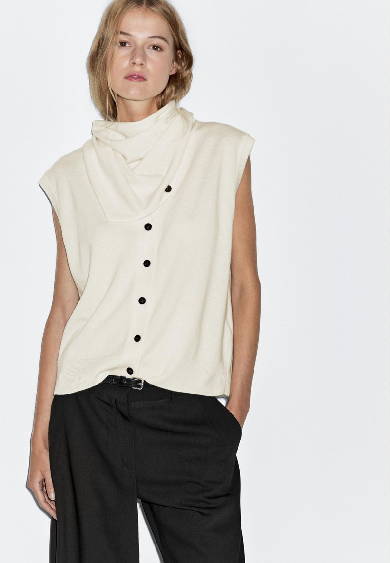 Beige ärmelloses Oberteil mit einem Cowl-Neck, das über eine Knopfleiste und schwarze Knöpfe verfügt. Kombiniert mit schwarzen weit geschnittenen Hosen.