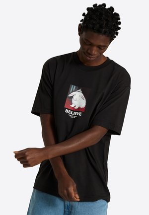 Ung mand iført en sort t-shirt med et billede af en hvid kanin og ordet "TROR" står mod en ensfarvet baggrund.