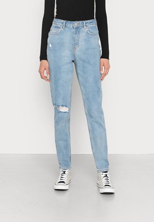 Personne portant un jean bleu clair déchiré, un haut noir à manches longues et des baskets montantes blanches à taches noires, debout sur un sol gris.
