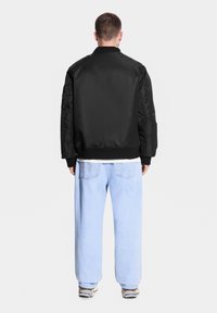 Czarna kurtka bomberka o gładkim wykończeniu, z prążkowanymi mankietami i luźnym krojem, zestawiona z jasnoniebieskimi dżinsami.