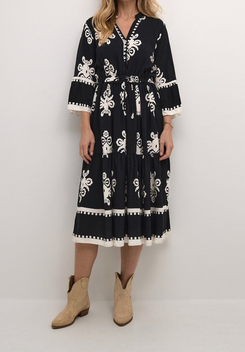 Femme portant une robe midi noire avec motifs floraux blancs, manches trois-quarts, et bottines beige, debout devant un fond uni.