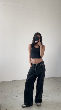 Jeans largos pretos de cintura alta, combinados com uma blusa justa preta em malha canelada. Ténis cinzentos com detalhes em branco. Fundo neutro.