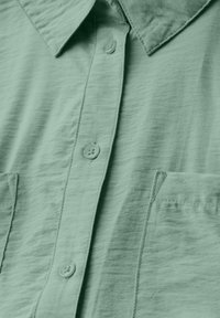 Camicia verde chiaro con collo, due tasche frontali e tessuto testurizzato con un aspetto leggermente stropicciato.
