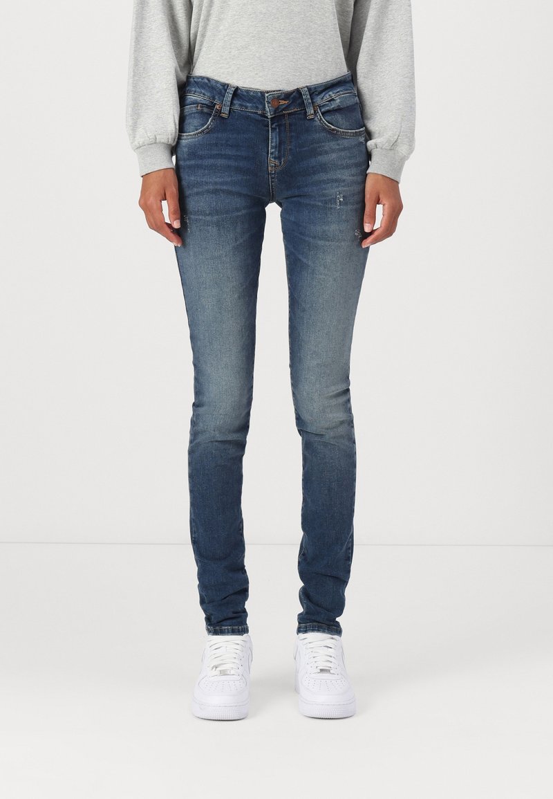 Vaqueros de denim azul ajustados con una cintura de altura media, ligeros desgastes y diseño de cinco bolsillos. Combinados con un sudadera gris y zapatillas blancas.