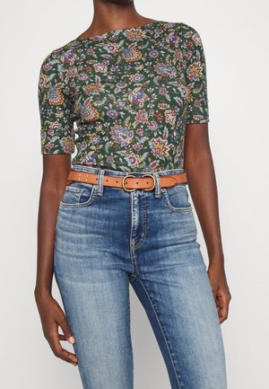 Femme portant un haut à manches courtes vert à motifs floraux, associé à un jean taille haute bleu clair et une ceinture en cuir beige.