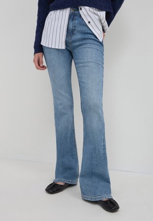 Person trägt hellblaue Schlagjeans, schwarze Ballerinas mit quadratischer Spitze, einen navyblauen Strickpullover und ein weißes Hemd mit schwarzen Streifen.