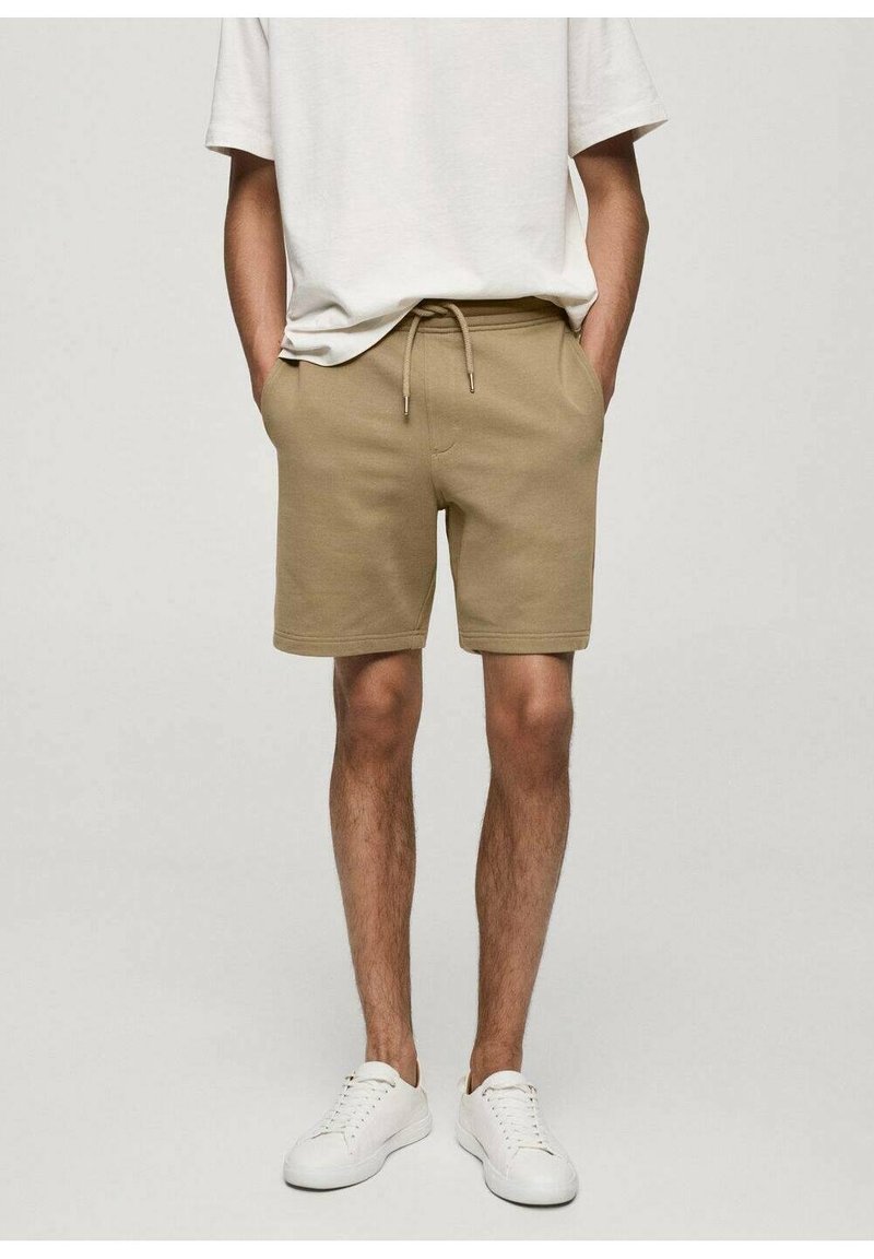 Shorts en coton beige avec une taille élastique et un cordon de serrage, dotés de deux poches latérales et d'une coupe décontractée, associés à des baskets blanches.