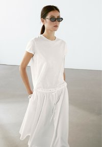Witte katoenen t-shirt met korte mouwen, gestoken in een vloeiende witte rok met een elastische tailleband en koord. Zwarte zonnebril.
