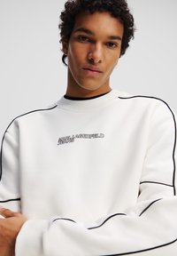 Sudadera blanca con detalles en negro, cuello acanalado y logo "KARL LAGERFELD JEANS" en el frente. Textura suave, corte relajado.