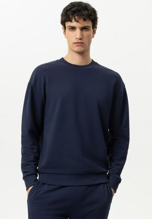 Giovane uomo con capelli corti e ricci, indossando una felpa blu scuro tinta unita e pantaloni abbinati, in piedi con le mani in tasca su sfondo bianco.