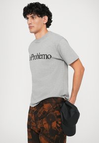 T-shirt grigia in cotone con testo "No Problemo" in nero, abbinata a pantaloni marroni con un motivo a stampa mista e un cappellino nero tenuto in mano.