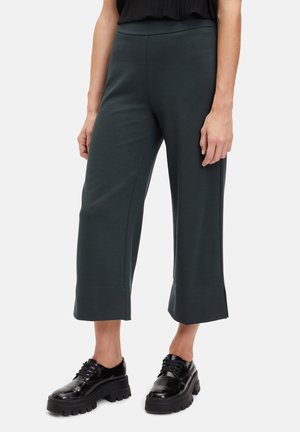 Pantalones recortados en verde oscuro, hechos de un material suave, con un diseño de pierna ancha y aberturas en los laterales del dobladillo, combinados con zapatos de plataforma negros.