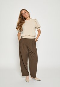 Béžový top s nadýchanými rukávmi v kombinácii s hnedými plisovanými nohavicami. Modelka má na sebe béžové topánky so špicatou špičkou. Hladká tkanina s jemnou textúrou.