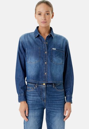 Camicia di jeans tagliata di colore blu medio con maniche lunghe, chiusura frontale con bottoni, due tasche sul petto e una discreta etichetta del marchio sulla tasca sinistra.