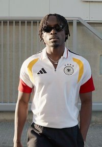 Férfi napszemüvegben és fehér, vörös és sárga részletekkel díszített német focimezben, szabadban, egy fémkerítés mellett áll.