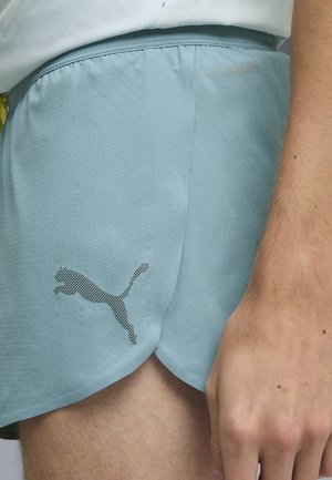 Pantaloncini sportivi azzurri chiari con logo Puma nero indossati sulla parte superiore della coscia di una persona e mano appoggiata accanto ai pantaloncini.