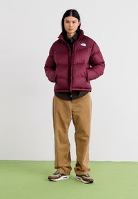 Burgunderröd pufferjacka med dragkedja fram, vadderad textur, bärs över en mörk skjorta och beige kordflöjlsbyxor. Sneakers har gröna och bruna accenter.