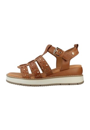 Sandalia plana de cuero marrón con múltiples correas con tachuelas, hebilla en el tobillo, suela acolchada y plataforma blanca en la suela.