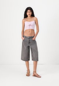 Lys rosa kort topp med front-ruching, kombinert med baggy grå denimshorts. Båret med brune sandaletter med stropper. Nøytral bakgrunn.