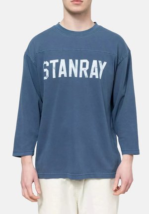 Personne portant une chemise bleue ample à manches longues avec un texte blanc "STANRAY" sur la poitrine, associée à un pantalon de couleur claire.
