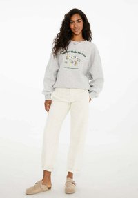 Sivá mikina s grafickým motívom ustrice a zeleným textom, v kombinácii s bielymi zužujúcimi sa nohavicami a béžovými dreváčkami. Mäkko tkanina a voľný strih.