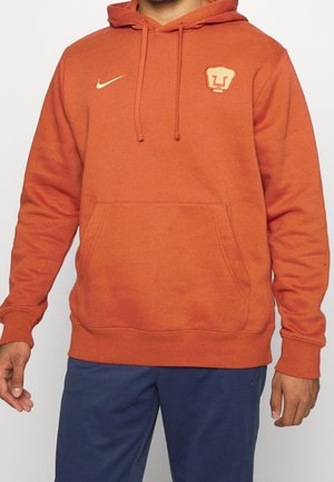 Homme portant un sweat à capuche orange avec poche avant, logo Nike sur le côté droit de la poitrine et emblème de visage d'animal sur le côté gauche de la poitrine, assorti à un pantalon bleu.