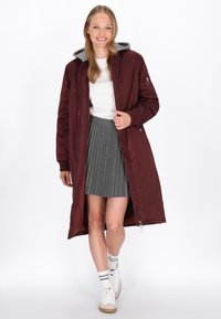 Abrigo largo burdeos con capucha gris, cierres de cremallera, puños acanalados y bolsillos. Lleva puesto un top blanco y una falda gris con plisados, junto con zapatillas blancas.