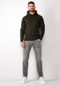 Felpa con cappuccio verde scuro e coulisse, abbinata a jeans slim-fit grigi chiari e sneakers bianche. Texture liscia, design casual.